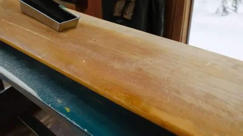 Un banco de trabajo de madera, ordenado y acogedor, está lleno de herramientas y un snowboard brillante, iluminado por la luz natural que entra por la ventana